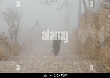 Un uomo anziano in un cappello e cappotto sale le scale nella nebbia sotto un ombrello nero. Uomo solitario triste in sciarpa con ombrello che cammina sotto la pioggia. Indietro Foto Stock