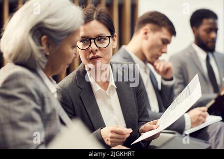 Attraente giovane donna d'affari in occhiali seduti in auditorium e parlare con il collega sul modulo di richiesta Foto Stock
