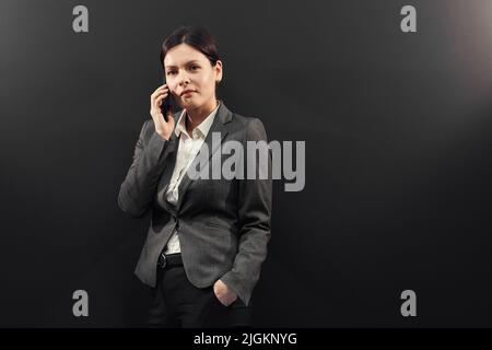 Ritratto di contenuti di successo giovane donna di vendita manager in giacca formale in piedi sfondo nero e parlare al telefono cellulare Foto Stock