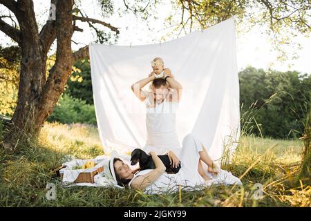 Giovane donna felice sorridendo sinceramente mentre si trova su coperta con il cane mentre il suo marito amorevole che porta la bambina sulle spalle. Bella famiglia caucasica di tre persone che si divertono all'aperto. Foto Stock