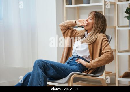 Sonnolenta stanca simpatica ragazza bionda in caldo maglione seduta sbadiglio e copre la sua bocca con mano in poltrona a casa. Pausa dal lavoro, fare una pausa Foto Stock