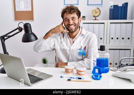 Giovane dentista ispanico uomo che lavora in clinica medica sorridendo facendo il gesto del telefono con le mani e le dita come parlare al telefono. Comunicando co Foto Stock