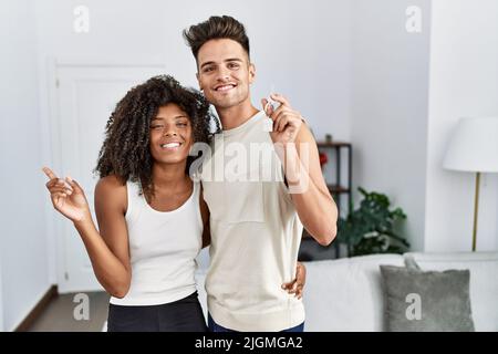 Giovane coppia interrazziale che tiene le chiavi della nuova casa sorridendo felice indicando con mano e dito al lato Foto Stock