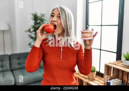 Donna di mezza età dai capelli grigi che bitting mela rossa e tiene la dentiera a casa Foto Stock