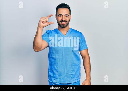 Bell'uomo ispanico con barba che indossa blu infermiere maschio uniforme sorridente e fiducioso gesturing con mano che fa segno di piccola taglia con le dita che guardano Foto Stock