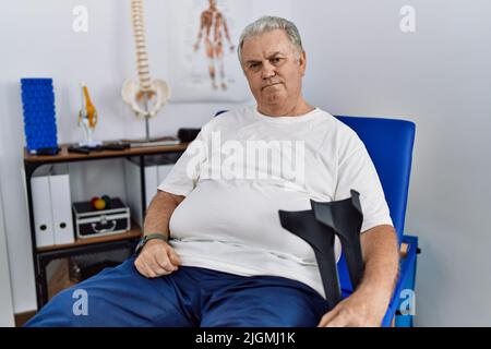 L'uomo caucasico anziano alla clinica di fisioterapia che tiene le stampelle che osservano assonnato e stanco, esaurito per affaticamento e hangover, occhi pigri alla mattina. Foto Stock