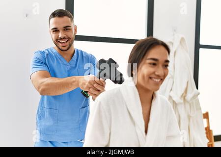 Latino uomo e donna che indossano la fisioterapia uniforme massaggiante collo con pistola a percussione al centro di bellezza Foto Stock