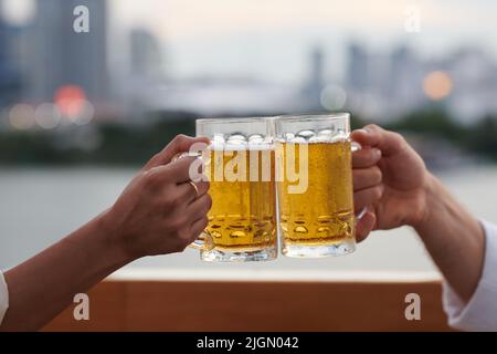 Immagine ritagliata di due persone che tostano con grandi tazze pesanti di birra fredda contro la vista della città Foto Stock