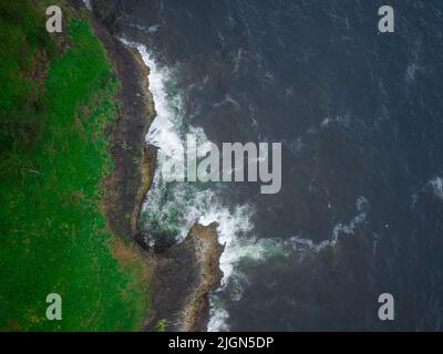 Una vista aerea di un oceano grigio scuro con onde bianche che si infrangono contro una tortuosa costa collinare coperta di lussureggiante erba verde. Paesaggio desertico, wil Foto Stock