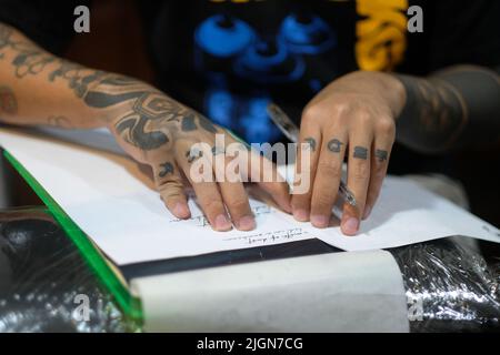 Primo piano delle dita inchinate del tatuatore mentre si ferma mentre stenciling la scritta cursive su carta bianca con una penna in preparazione per una sessione di tatuaggio. Foto Stock