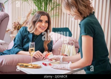 Donna allegra che punta al campione di unghie di colore rosso e sorridente mentre sceglie manicure con manicurista. Manicure master in guanti sterili che reggono unghie lucidare tavolozza di colori. Foto Stock