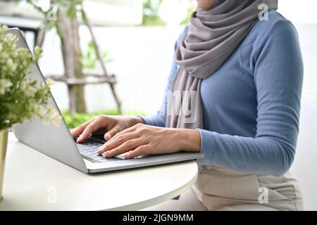 Immagine ritagliata, giovane musulmana asiatica che indossa hijab, seduto nel cortile della casa e utilizzando un computer portatile. Foto Stock