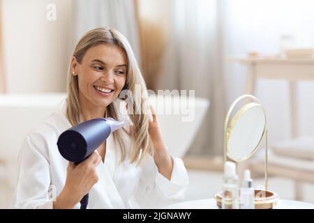 Bella signora bionda di mezza età usando l'asciugacapelli a casa Foto Stock