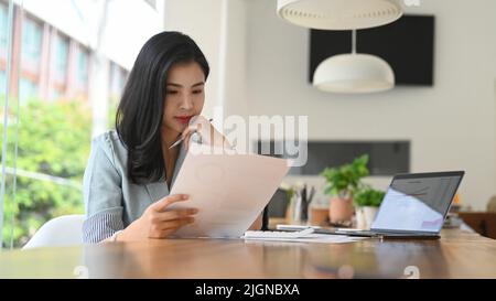 Seria giovane donna d'affari che legge il documento di statistiche, progettando la strategia di affari alla scrivania d'ufficio Foto Stock