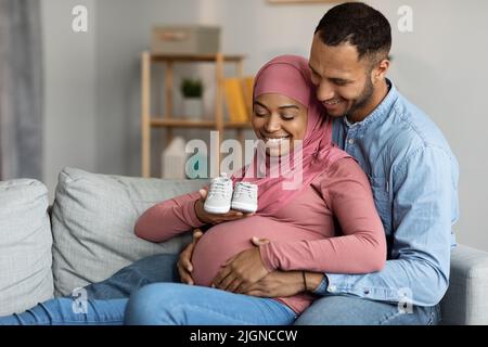 Happy Black Muse musulmane che tiene piccole scarpe per bambini mentre si rilassa sul divano Foto Stock
