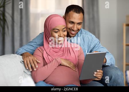 Buone spose musulmane nere incinte che usano il Tablet digitale mentre si rilassano a casa Foto Stock