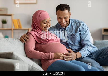 Ritratto di felice coppia islamica nera in attesa di bambino che riposa insieme sul divano Foto Stock