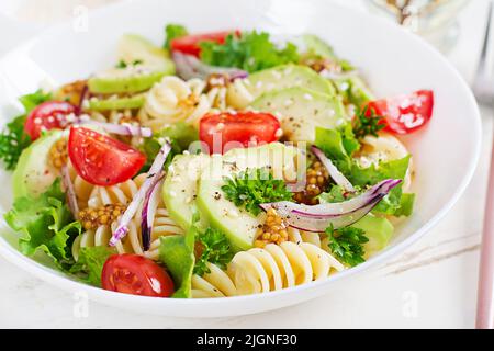 Insalata di pasta fusilli con avocado, pomodori, lattuga fresca verde, cipolla rossa e senape condita su sfondo bianco. Pranzo vegetariano sano. Foto Stock