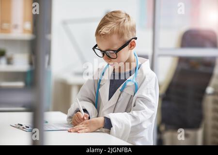 Medico serio occupato poco in occhiali e cappotto bianco in piedi a tavola in ufficio medico e riempire scheda medica Foto Stock