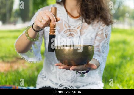 Dettaglio delle mani di una donna che tiene e suona una ciotola di canto tibetano durante una sessione di meditazione e terapia musicale in un parco. Foto Stock