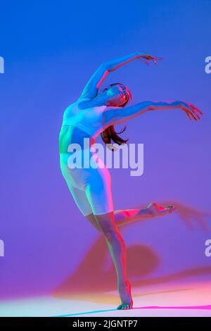 Giovane ragazza flessibile grazioso in sport fitness uniforme danza isolata su sfondo rosa-viola sfumato in luce al neon. Sport, bellezza, pubblicità Foto Stock