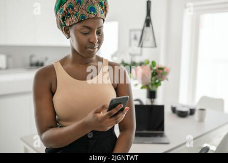 Bella donna africana nera che lavora a casa, sorridente, usando smartphone mobile, indossando il foulard tradizionale Foto Stock