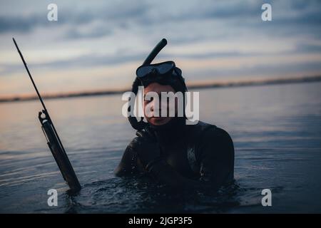 Buon subacqueo giovane che guarda la macchina fotografica mentre tiene una pistola e indossa un equipaggiamento da scuba. Giovane uomo che pesca in mezzo al mare. Foto Stock