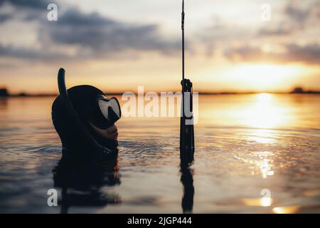 Maschio subacqueo che tiene una pistola mentre immerso in acqua di mare nel mezzo dell'oceano. Subacqueo a caccia di pesce al tramonto. Foto Stock