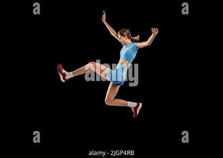 Giovane ragazza sportiva, lungo jumper in uniforme blu sportivo esegue il triplo salto isolato su sfondo nero. Concetto di sport, energia, realizzazioni Foto Stock