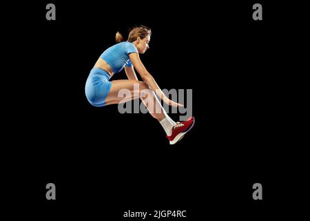 Giovane ragazza sportiva, lungo jumper in uniforme blu sportivo esegue il triplo salto isolato su sfondo nero. Concetto di sport, energia, realizzazioni Foto Stock