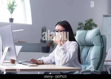 Sensazione di malessere al workplace.A giovane donna asiatica d'affari, freelancer, manager, dipendente si ammalò. È seduto alla scrivania strofinando il naso con un tovagliolo, ha un naso che cola, e si sente male. Foto Stock