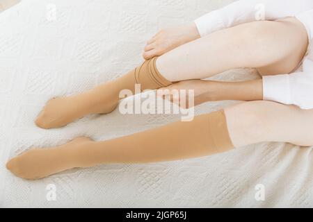 Gambaletti o calzini. Calze a compressione beige su una donna in una stanza bianca. Ragazza che mette su calze a casa. Belle gambe femminili Foto Stock