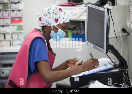 Un infermiere del sistema NHS in scrub controlla le registrazioni dei pazienti in una sala operatoria durante un intervento. Foto Stock