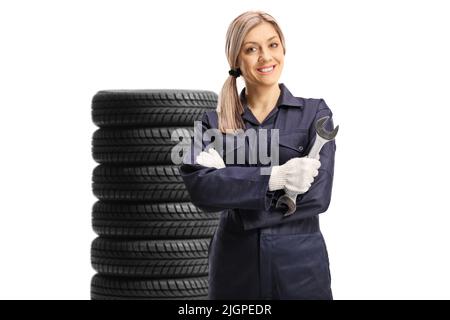 Giovane donna meccanica che tiene una chiave davanti ad un mucchio di pneumatici isolati su sfondo bianco Foto Stock