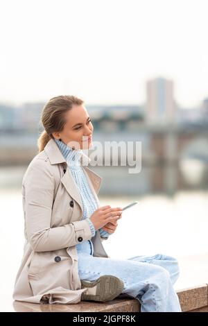Una giovane donna con smartphone seduta sul terrapieno in una giornata di primavera. Sorride e guarda via. Foto Stock