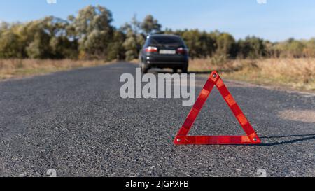 Triangolo segnaletico rosso sulla strada di fronte a un'auto rotta. Guasto della vettura in tempo di sole. Sicurezza del traffico stradale Foto Stock