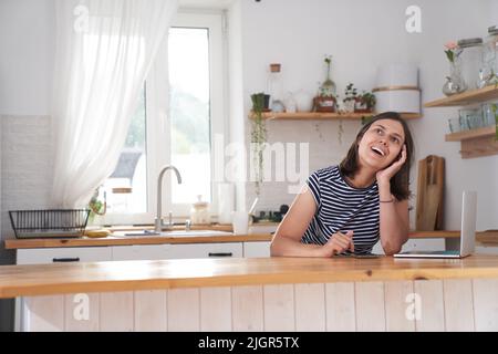 Una donna felice è in cucina nella sua casa. La donna ride felicemente e guarda via. Accanto a lei c'è un notebook. Il concetto di lavoro a distanza. Foto di alta qualità Foto Stock