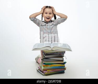 piccola ragazza affascinante con pigtail intrecciato siede di fronte a una grande pila di libri e tiene la testa con le mani in sorpresa. Foto Stock