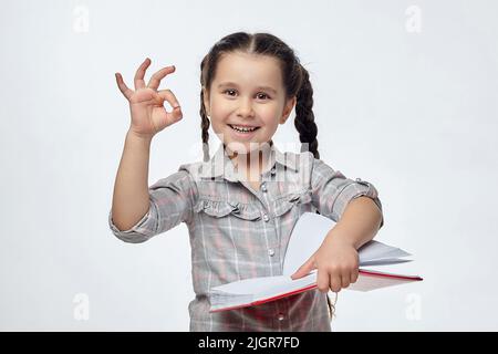 La bambina tiene un libro rosso nelle sue mani e mostra il segno OK. Foto Stock