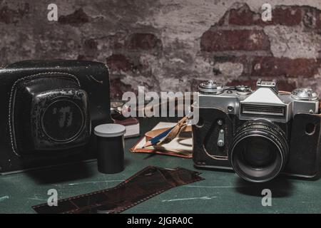 In cima a un tavolo usurato e graffiato vi è una vecchia e sporca fotocamera analogica, negativi fotografici, un contenitore di pellicola, la custodia della fotocamera, un notebook, una matita Foto Stock