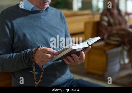 Mani di uomo anziano che tengono aperta la Bibbia e perle di rosario di legno mentre leggono i versi del Vangelo o di qualche altro libro durante il sermone Foto Stock