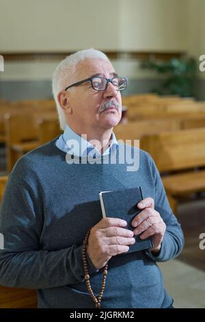 Uomo serio invecchiato con capelli grigi che tengono la Sacra Bibbia e perle di rosario di legno dal petto mentre si concentra sulla preghiera mentre si siede in chiesa Foto Stock