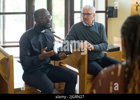 Pastore fiducioso in camicia nera con colletto clericale che spiega i versi dalla Bibbia ai parrocchiani interculturali della sua chiesa dopo il sermone Foto Stock