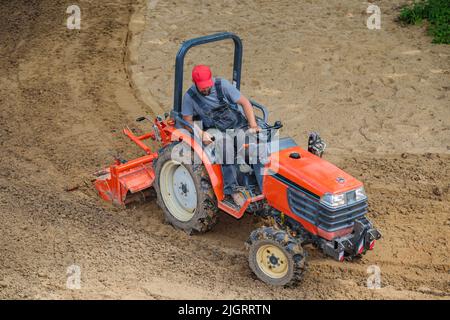 Un agricoltore su un mini trattore allenta il terreno per il prato. Coltivazione di terra, livellamento di superficie. Foto Stock