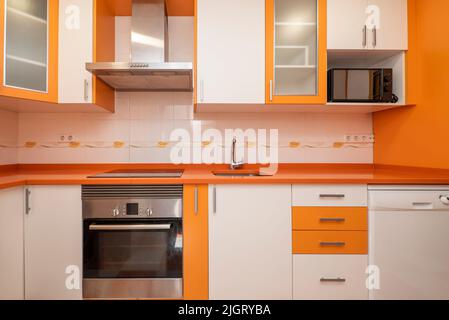 Cucina con mobili bianchi e arancioni luminosi, cassetti e forni, piano di lavoro in pietra arancione, accenti grigi e elettrodomestici incorporati Foto Stock