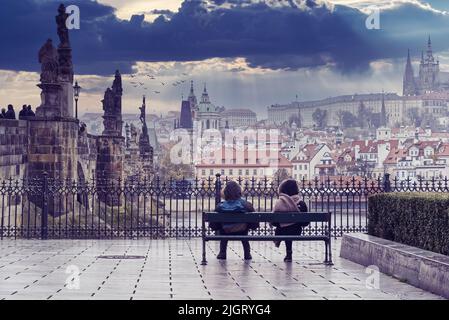 Un paio di giovani seduti sulla panchina guardando l'Hradcany di Praga con una vista parziale del ponte Carlo e del fiume Moldava Foto Stock