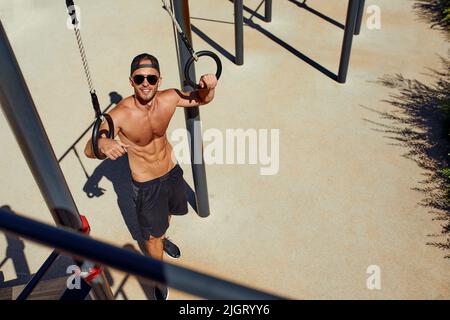 Sportivo muscoloso che si esercita su anelli ginnici all'aperto in palestra all'aperto. Mani agli anelli che immergere l'uomo facendo l'esercitazione usando gli anelli Foto Stock