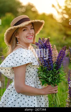 una giovane donna con un bouquet di lupini in un abito a pois polka e un cappello di paglia cammina sul prato con lupino al tramonto Foto Stock