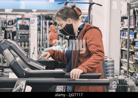 Donna che indossa occhiali e una maschera contro il virus acquista un grande tapis roulant in un negozio Foto Stock