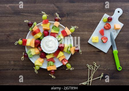 Raffigurazione della preparazione di un piatto di spiedini di frutta fresca, su un tavolo di legno scuro. Foto Stock